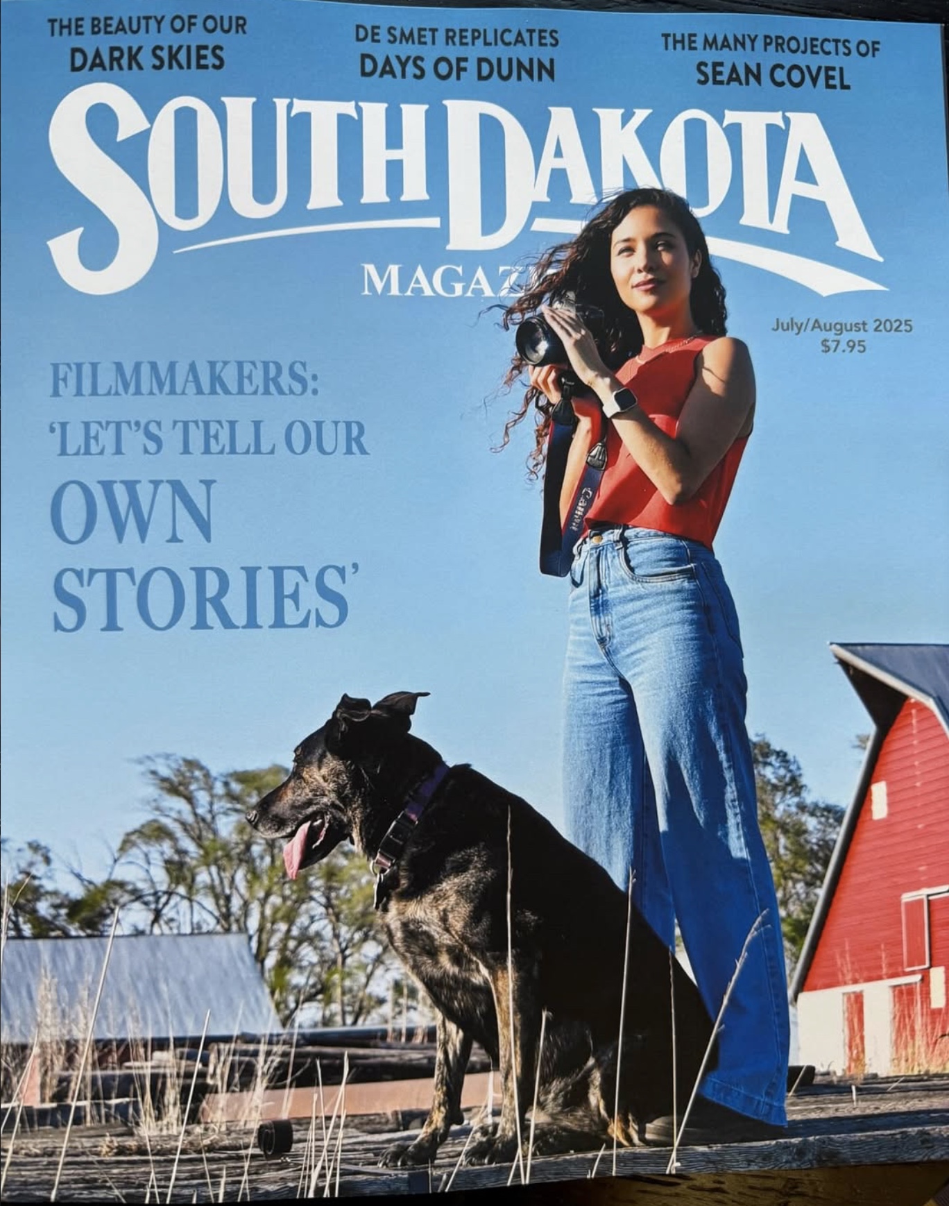 Charlie Buhler holding a camera beside a dog on the July/August 2025 cover of South Dakota Magazine.
