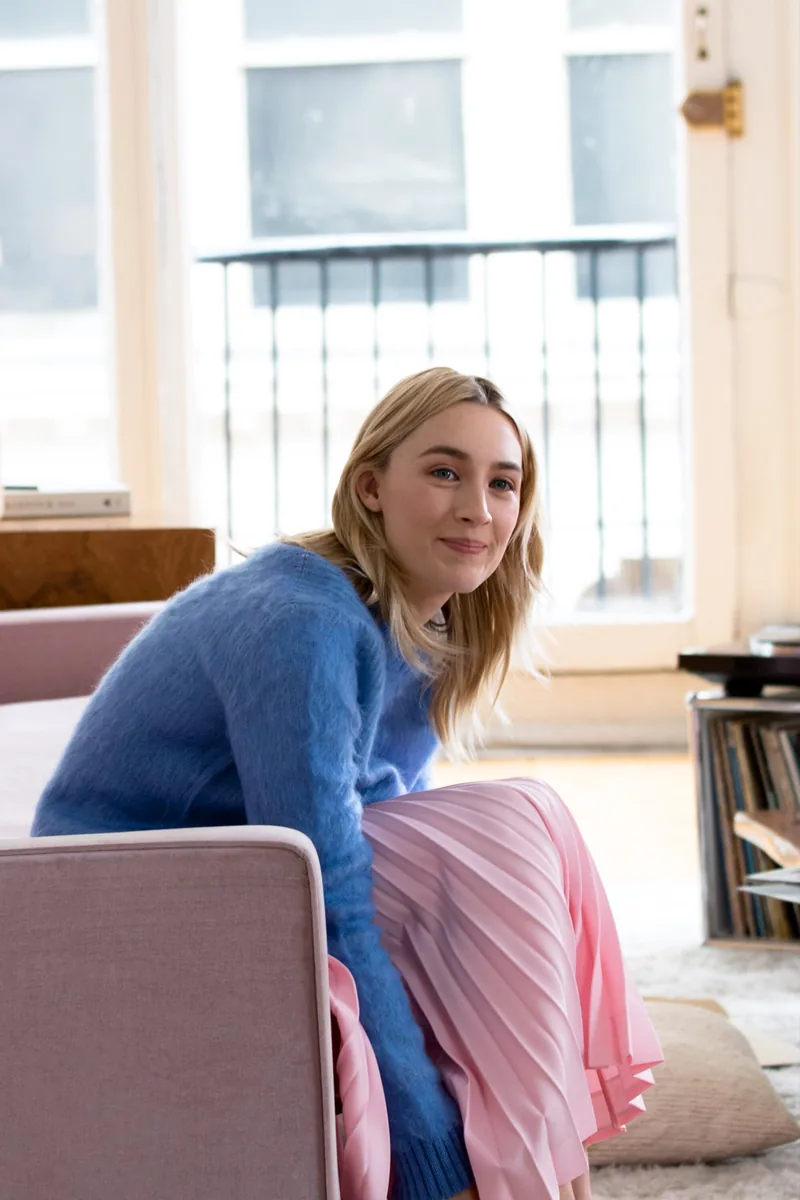 A model in a blue sweater and pink skirt seated beside a bright window.
