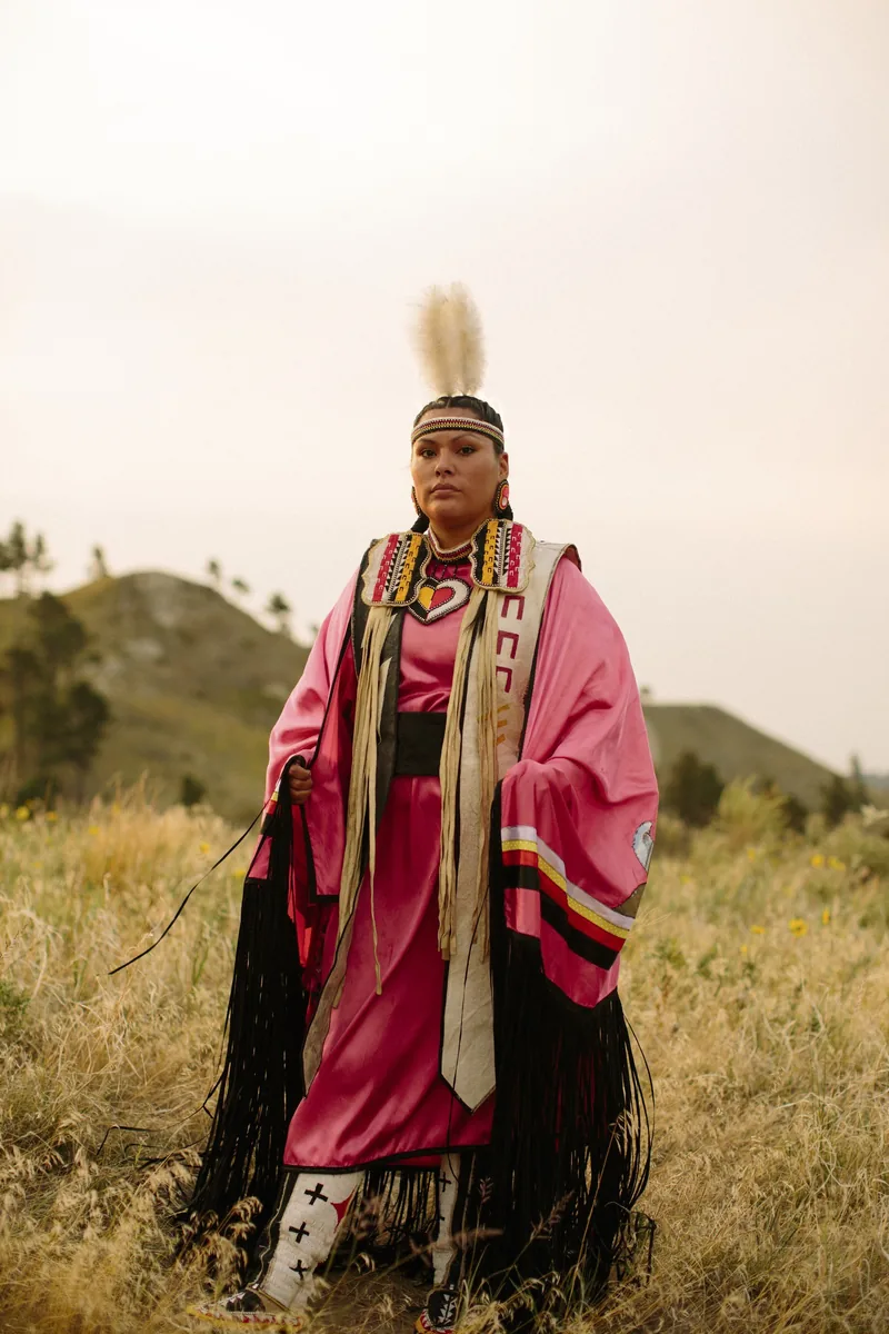 A person wearing pink fringed regalia standing in a grassy field.
